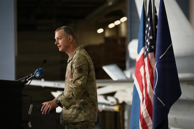 General håller tal vid ceremonipodium utomhus på flygbas, allierade militärpersoner i bakgrunden.